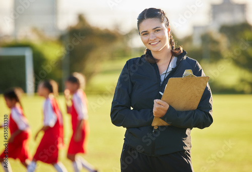 Soccer field, woman coach with and girl team training on grass in background. Sports, youth development and teamwork, a happy young female volunteer coaching football team with clipboard from Brazil.