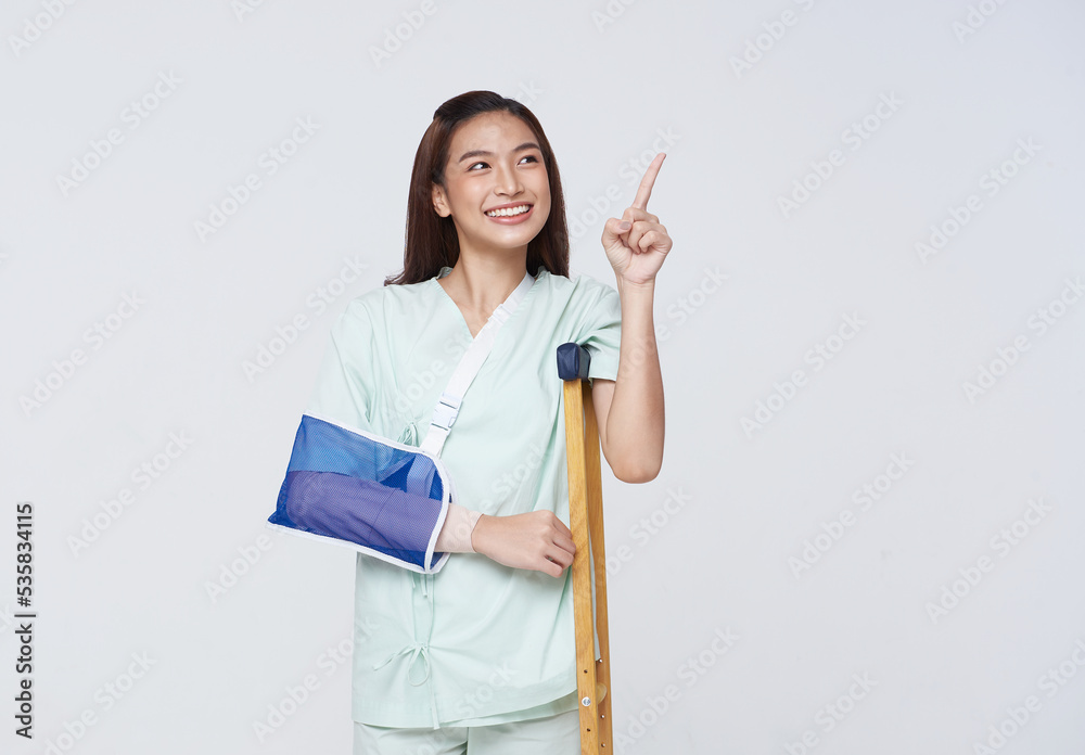 Asian teenage woman wearing a patient gown put on a cast Use a crutch to walk due to accidental injury standing pointing to empty space with awesome smile insurance concept. Asian teenage woman wearing a patient gown put on a cast Use a crutch to walk due to accidental injury standing pointing to empty space with awesome smile insurance concept.