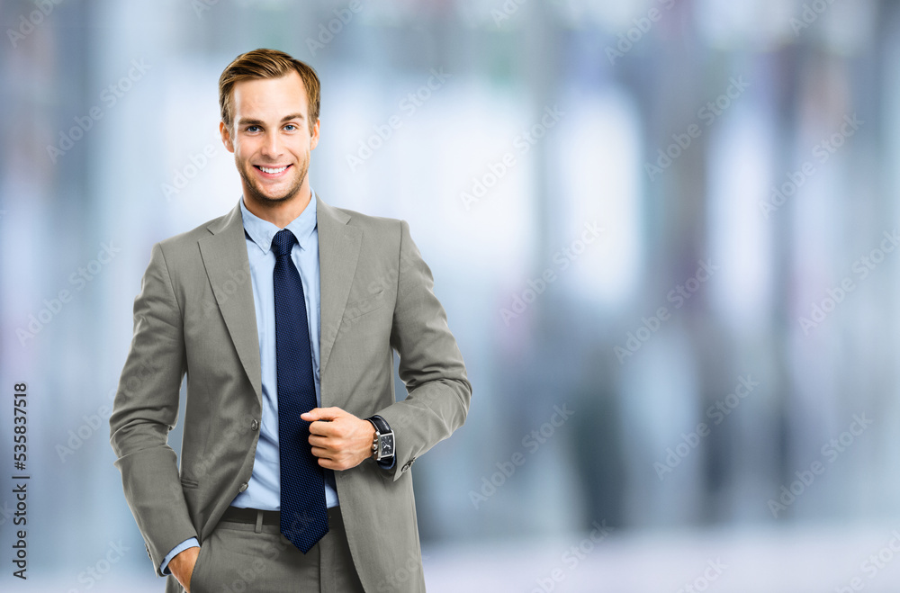Portrait of happy confident businessman in grey suit, blue shirt and tie, over blurred office background. Business success concept. Smiling man at studio picture. Portrait of happy confident businessman in grey suit, blue shirt and tie, over blurred office background. Business success concept. Smiling man at studio picture.