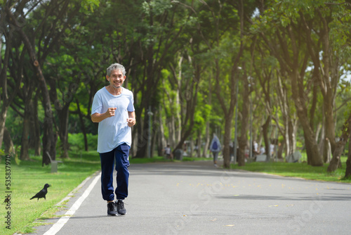 senior man with grey hair and beard wearing headphones for listening music is running in the park, soft focus.concept elderly people lifestyle, workout,healthcare, happiness, wellness