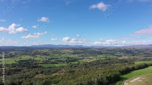 Aerial shot flying down to a rocky cliff face with countryside meadows, mountains and rural villages in the background. Filmed in the Lake District, UK on a sunny summer day. photo