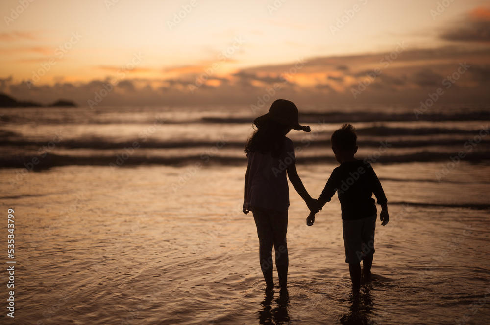 Silhouette portrait of sibling holding hands on the beach at sunset.