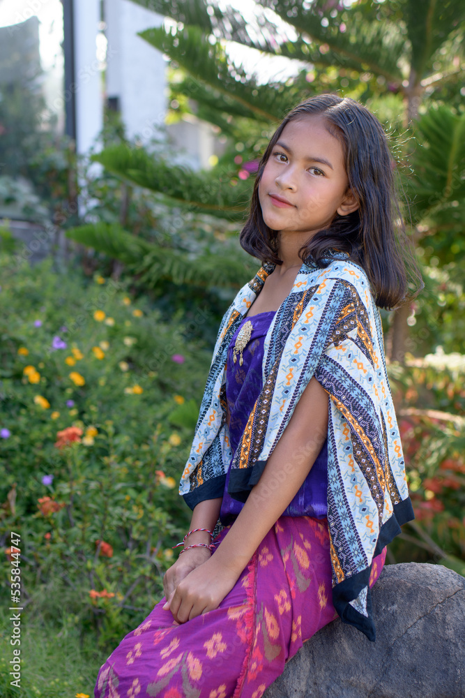 Young Bali girl in traditional suit sarong poses in colorful in garden with flowers. Young Bali girl in traditional suit sarong poses in colorful in garden with flowers.