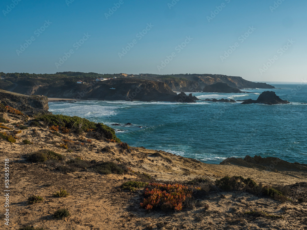 View of Zambujeira do Mar over the sea shore with ocean waves, cliffs and sand dunes covered by green vegetation red leaves of sour fig, sunny day, clear blue sky. Rota Vicentina coast, Portugal View of Zambujeira do Mar over the sea shore with ocean waves, cliffs and sand dunes covered by green vegetation red leaves of sour fig, sunny day, clear blue sky. Rota Vicentina coast, Portugal
