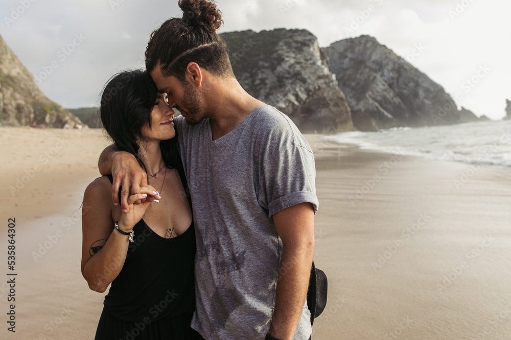 joyful bearded man and tattooed woman in dress holding hands near ocean.