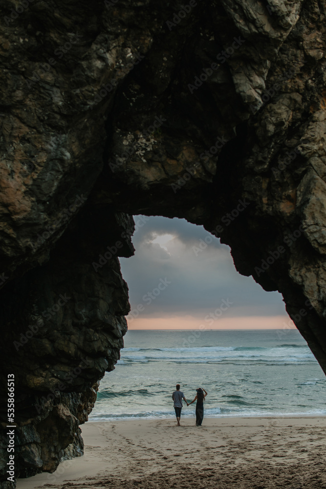 Fototapeta premium back view of couple holding hands while standing on beach during sunset.