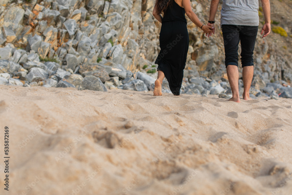 cropped view of couple holding hands and walking on sand near rocks. cropped view of couple holding hands and walking on sand near rocks.
