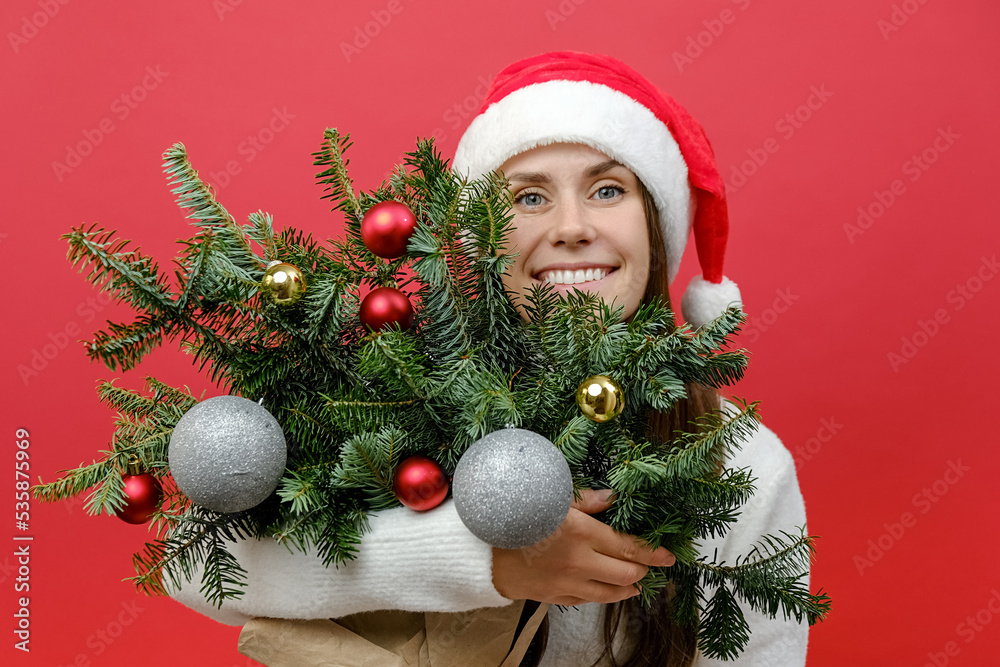 Portrait of cheerful beautiful young woman wears white sweater and Santa hat holding hug bouquet of spruce branches look up, isolated on red background. Happy New Year 2023 celebration holiday concept Portrait of cheerful beautiful young woman wears white sweater and Santa hat holding hug bouquet of spruce branches look up, isolated on red background. Happy New Year 2023 celebration holiday concept