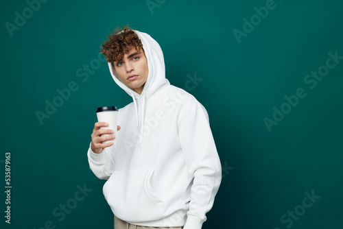 a handsome man stands on a green background in a white hoodie holding a glass in his hand and looks at the camera. Horizontal Studio Photography © Tatiana