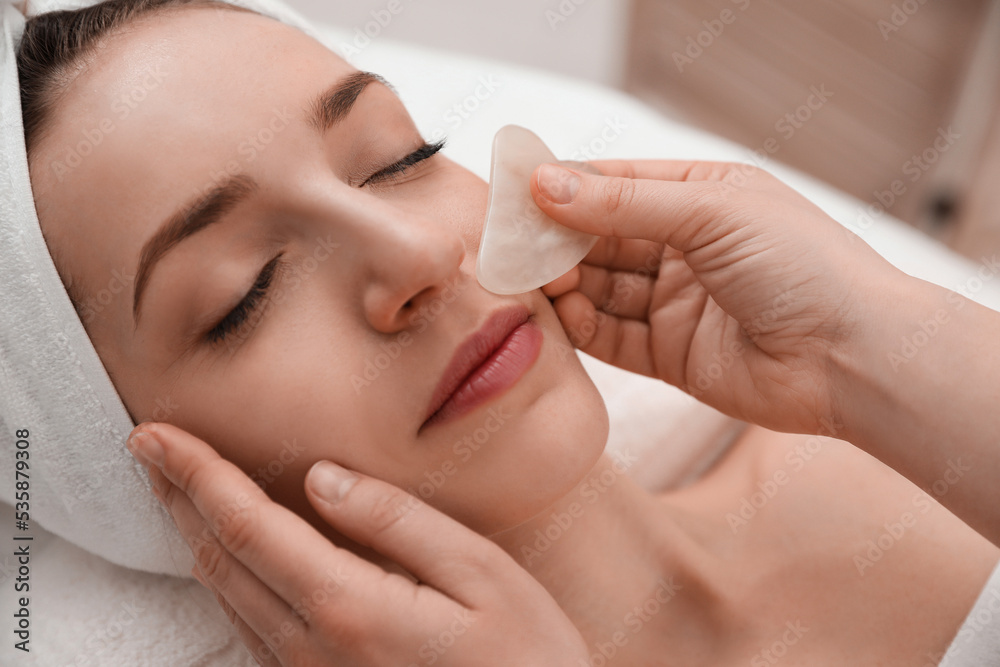 Young woman receiving facial massage with gua sha tool in beauty salon, closeup
