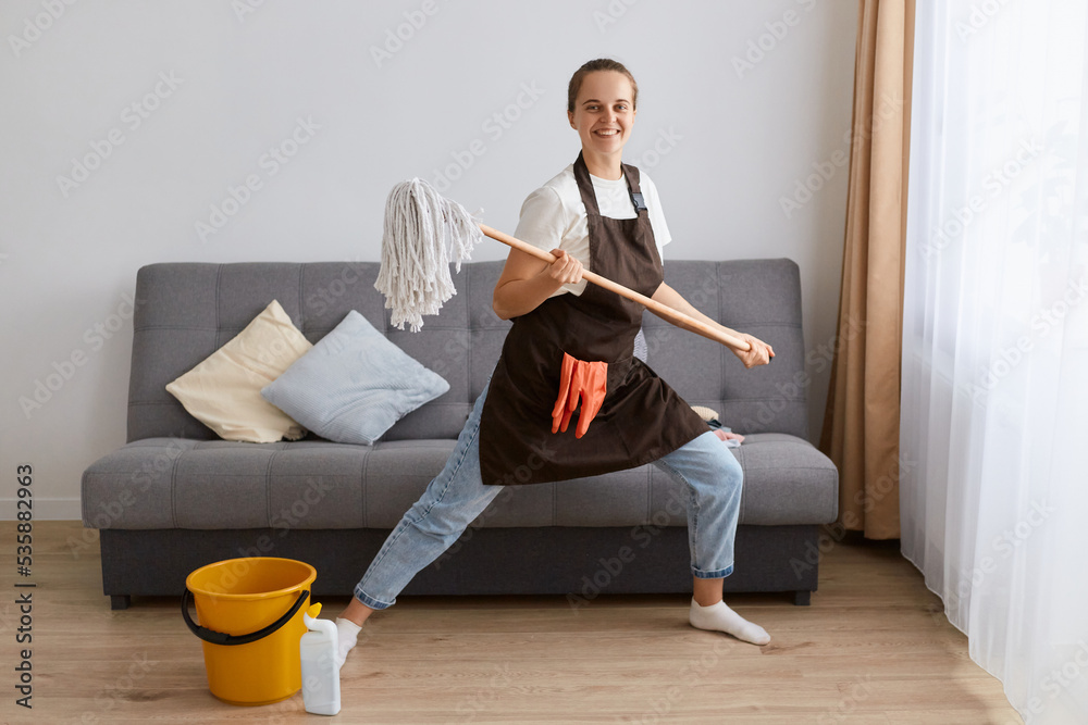 Portrait of funny optimistic female washing floor with mop at home, doing domestic chores and having fun, dancing with cleaning tool, expressing positive emotions.