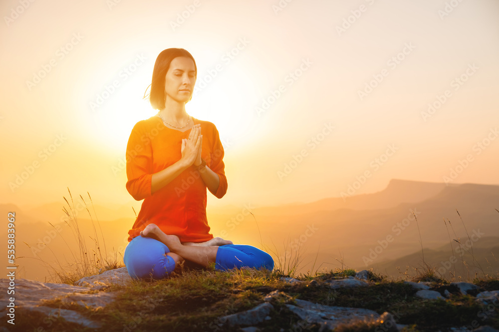Yogini in the lotus position in full face sits on a cliff against the backdrop of the sunset sky with mountains, prays