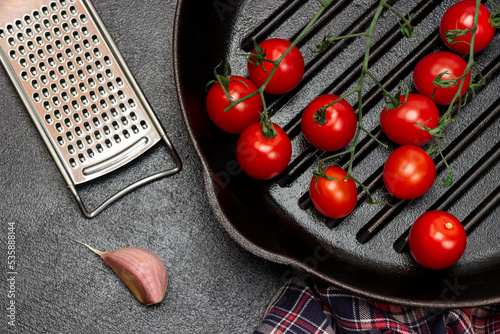 A sprig of cherry tomato in frying pan. Grater and cloves of garlic on table.