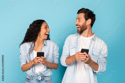 Surprised happy excited young diverse couple with smartphones look at each other, rejoice online victory