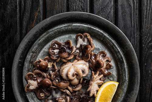 A dish of cooked baby octopus with a slice of lemon, shot from above, dark wooden backdrop