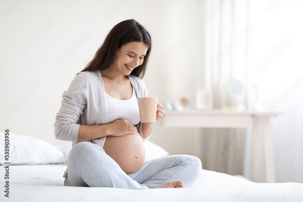 Smiling pregnant woman sitting on bed, drinking tea Smiling pregnant woman sitting on bed, drinking tea