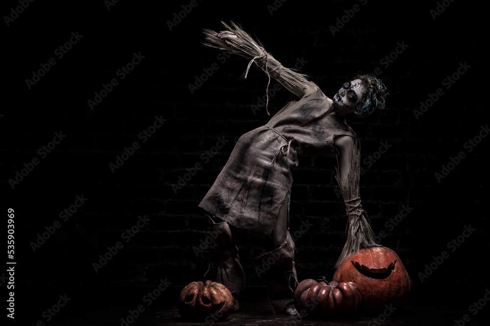 Scarecrow girl is standing near pumpkins at the dark background