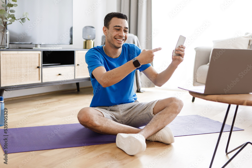 Happy arab male fitness blogger showing cellphone to laptop webcamera, recommending new app for training Happy arab male fitness blogger showing cellphone to laptop webcamera, recommending new app for training