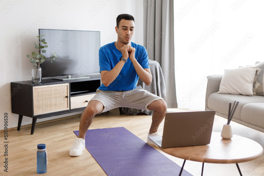 Young arab athlete man in sports clothes squatting in living room interior, watching online fitness class on laptop Young arab athlete man in sports clothes squatting in living room interior, watching online fitness class on laptop