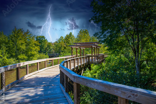 a long winding brown wooden bridge with a pergola over the Sandy Run Creek surrounded by lush green trees and plants with storm clouds and lightning at The Walk at Sandy Run in Warner Robins Georgia photo