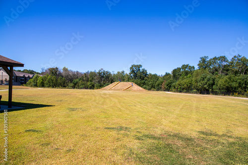 a large mound with two yellow and blue slides down the side surrounded by lush green grass and trees with a gorgeous clear blue sky at The Walk at Sandy Run in Warner Robins Georgia USA photo