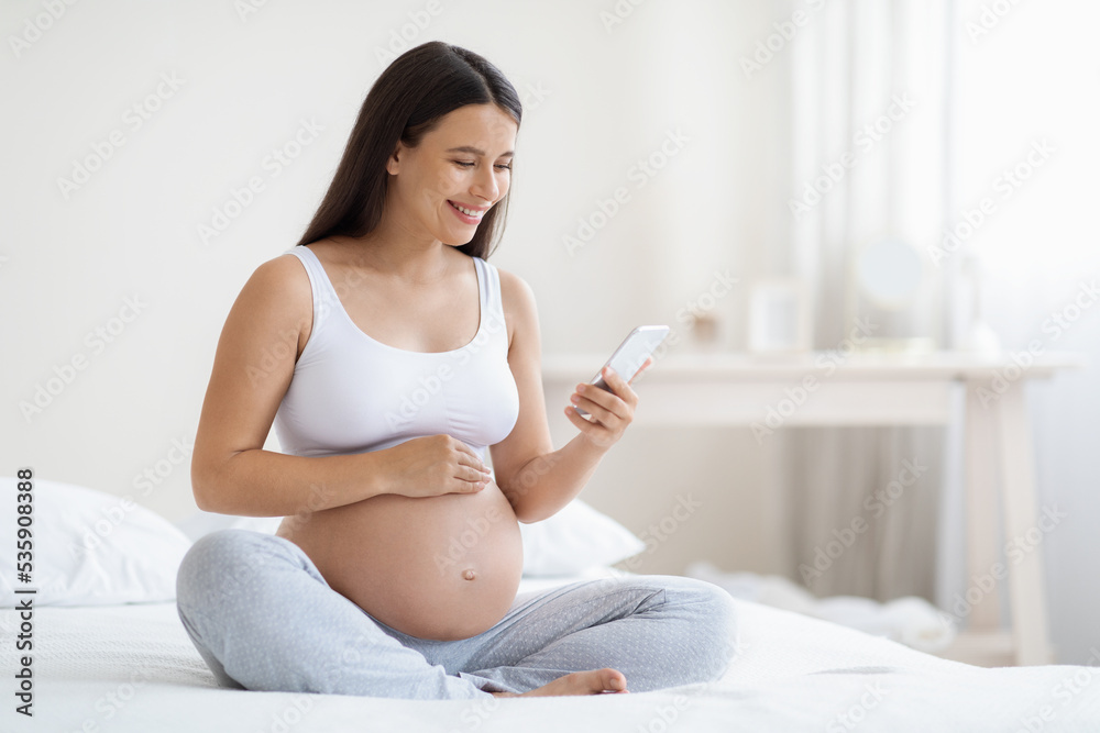 Happy expecting lady using smartphone, bedroom interior Happy expecting lady using smartphone, bedroom interior