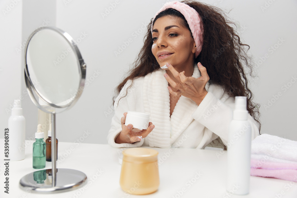 Smiling tanned pretty curly Latin lady apply facial cream admiring herself in mirror doing beauty procedure in white home interior. Copy space. Aesthetic medicine skincare beauty products ad concept Smiling tanned pretty curly Latin lady apply facial cream admiring herself in mirror doing beauty procedure in white home interior. Copy space. Aesthetic medicine skincare beauty products ad concept