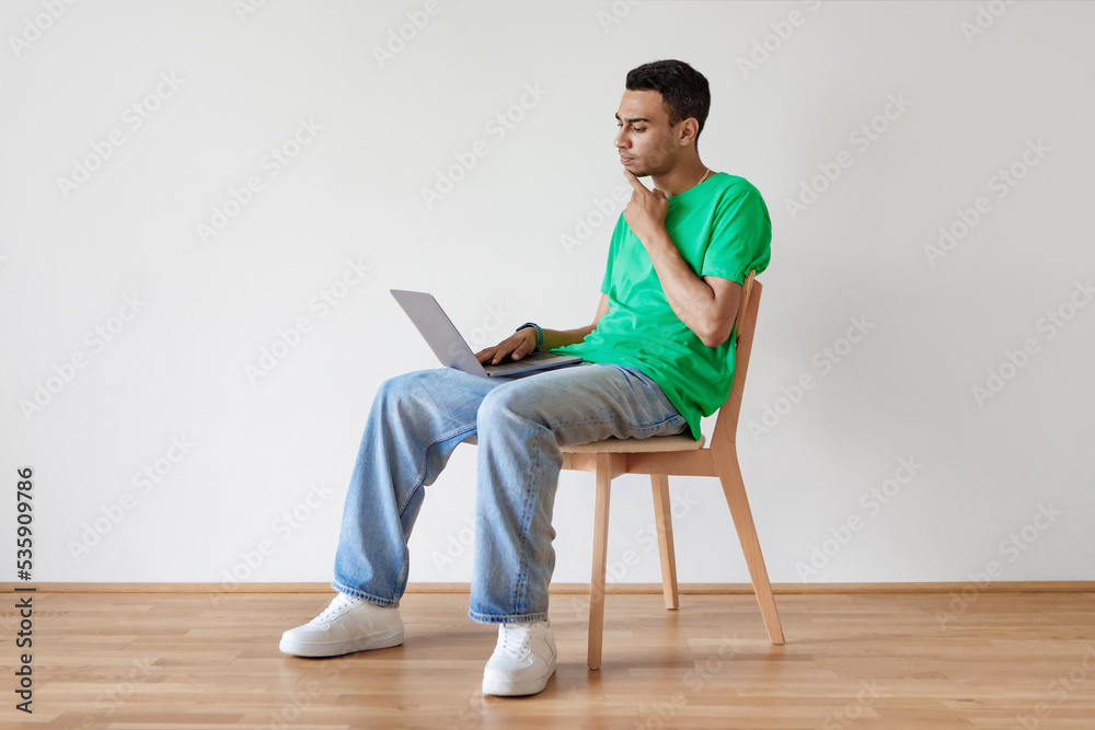 Troubled arab man sitting on chair with laptop, thinking over solution to problem against white wall, copy space