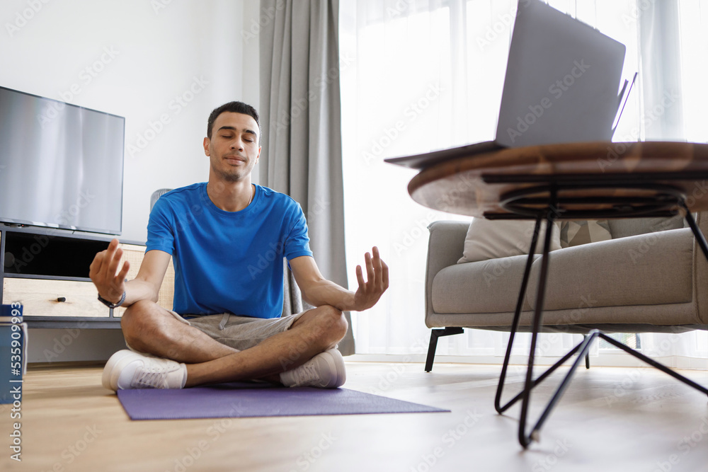 Domestic yoga. Arab man meditating in front of laptop at home, sitting with closed eyes in lotus position Domestic yoga. Arab man meditating in front of laptop at home, sitting with closed eyes in lotus position