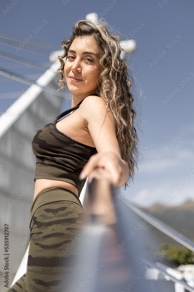 posing in a sculpture of the city a woman with curly and long hair, wears sportswear with a top and lycra with a military print, photo outside in the day