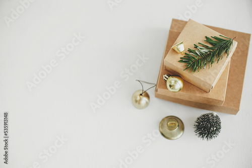 Stylish eco christmas gift boxes on white table flat lay. Simple craft christmas presents with golden baubles, bell and fir branch on rustic wooden table. Space for text. Merry Christmas