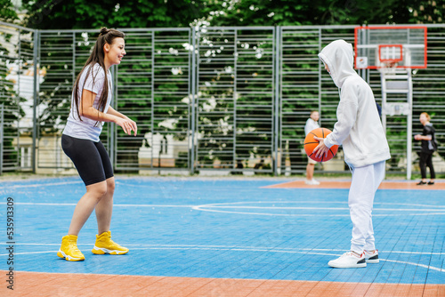 Girl and her younger brother, teenager, play basketball on modern basketball court under open sky