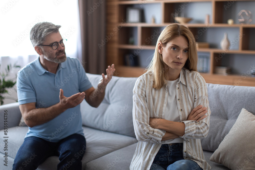 Angry mature caucasian man scolds to female on couch, quarreling in living room interior Angry mature caucasian man scolds to female on couch, quarreling in living room interior