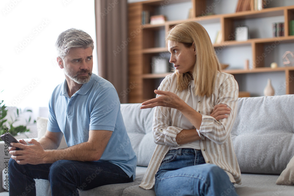 Offended angry mature caucasian wife scolds to husband with phone for gambling and gadget addiction on couch Offended angry mature caucasian wife scolds to husband with phone for gambling and gadget addiction on couch