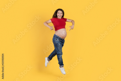Joyful Young Pregnant Woman Pointing At Her Belly While Jumping In Air