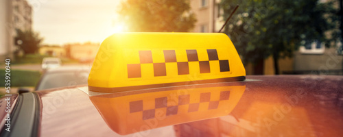 Fotografering Yellow checkered taxi sign fixed on cab roof waiting for passenger at urban stre