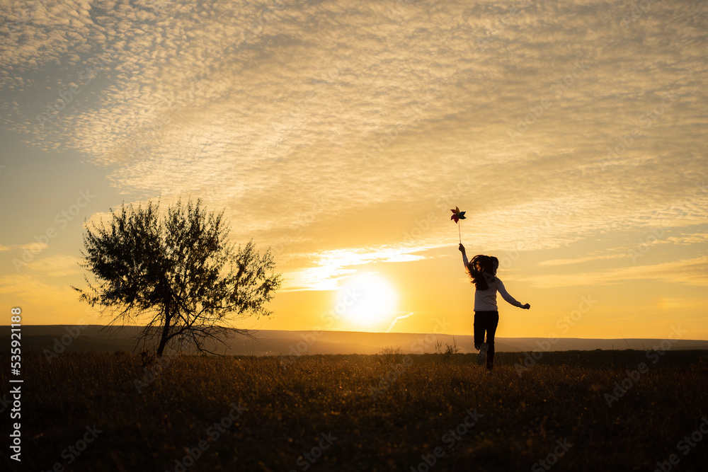 Little girl running on the meadow at sunset with windmill in her hands. Silhouette of child girl holding wind toy Little girl running on the meadow at sunset with windmill in her hands. Silhouette of child girl holding wind toy