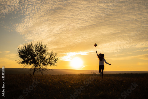 Little girl running on the meadow at sunset with windmill in her hands. Silhouette of child girl holding wind toy