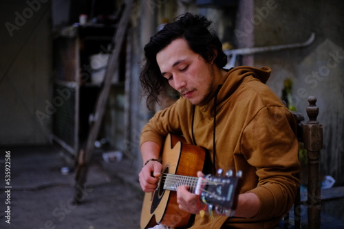 One Asian guy with medium length black curly hair sits outside on a wooden chair and plays the guitar.