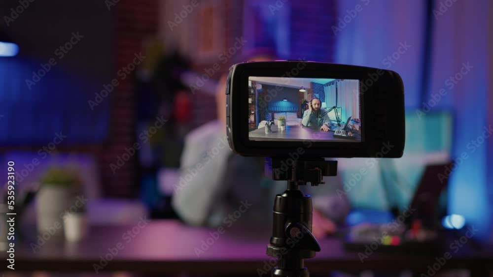 Selective focus on digital video camera screen recording male vlogger with headphones talking at microphone in home podcasting studio. Closeup of dslr filming influencer streaming live internet show. Selective focus on digital video camera screen recording male vlogger with headphones talking at microphone in home podcasting studio. Closeup of dslr filming influencer streaming live internet show.