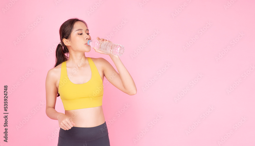 Sporty asian woman drinking fresh pure water in bottle after workout standing on pink background. Slim fit body young girl wear sportswear drink fresh water for healthy over isolated pastel background Sporty asian woman drinking fresh pure water in bottle after workout standing on pink background. Slim fit body young girl wear sportswear drink fresh water for healthy over isolated pastel background