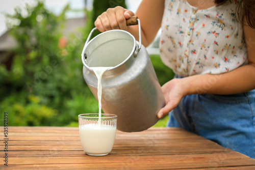 Woman pouring fresh milk from can into glass at wooden table outdoors, closeup