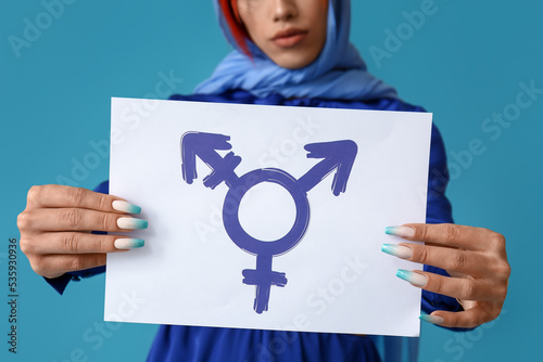 Stylish transgender woman holding paper sheet with transgender symbol against blue background, closeup photo