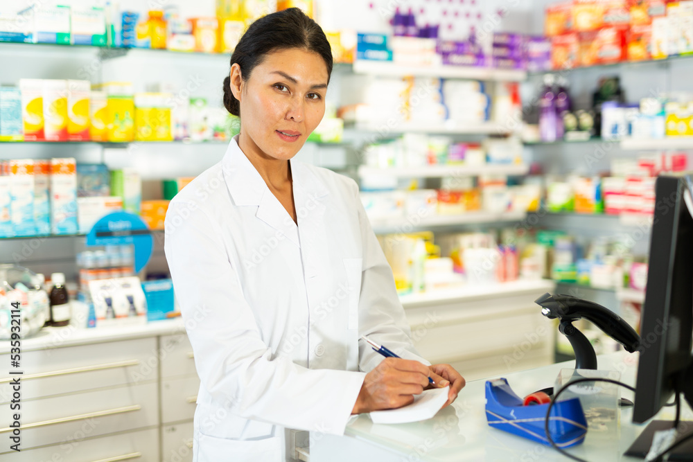 Asian woman doctor standing at counter in drugstore and writing recipe.