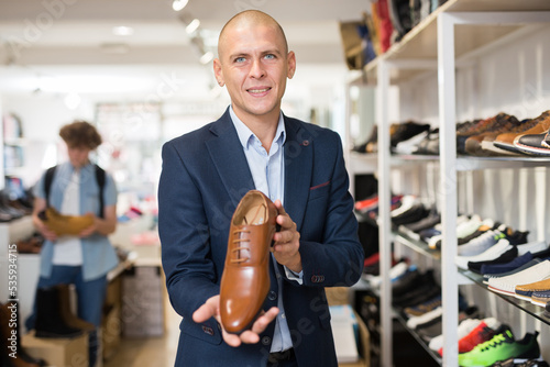 Friendly smiling salesman in blue suit demonstrating stylish brown leather model of mens formal shoes while standing near shelves in shoe boutique photo