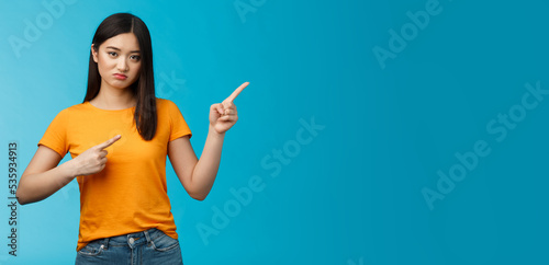 Picky disappointed asian girl with unsatisfied face, pointing upper right corner, sulking offended, feel jealous friend went summer vacation abroad alone, stand blue background displeased