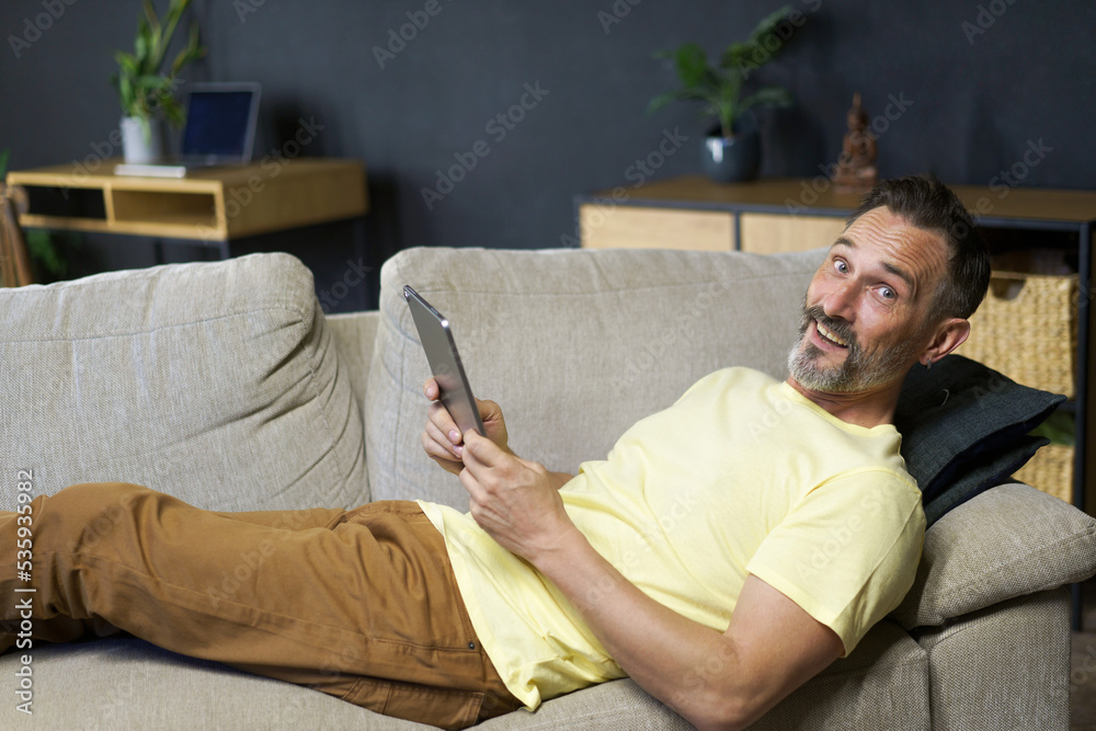 Laying on the couch middle aged freelancer man use digital tablet to read social media or working offline from home wearing casual. Relaxed freelancer man laying on sofa making business uses gadget