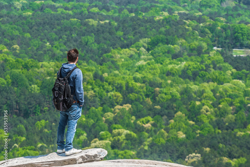 A young man with a backpack standing on the edge of the cliff and looking at the green forest below