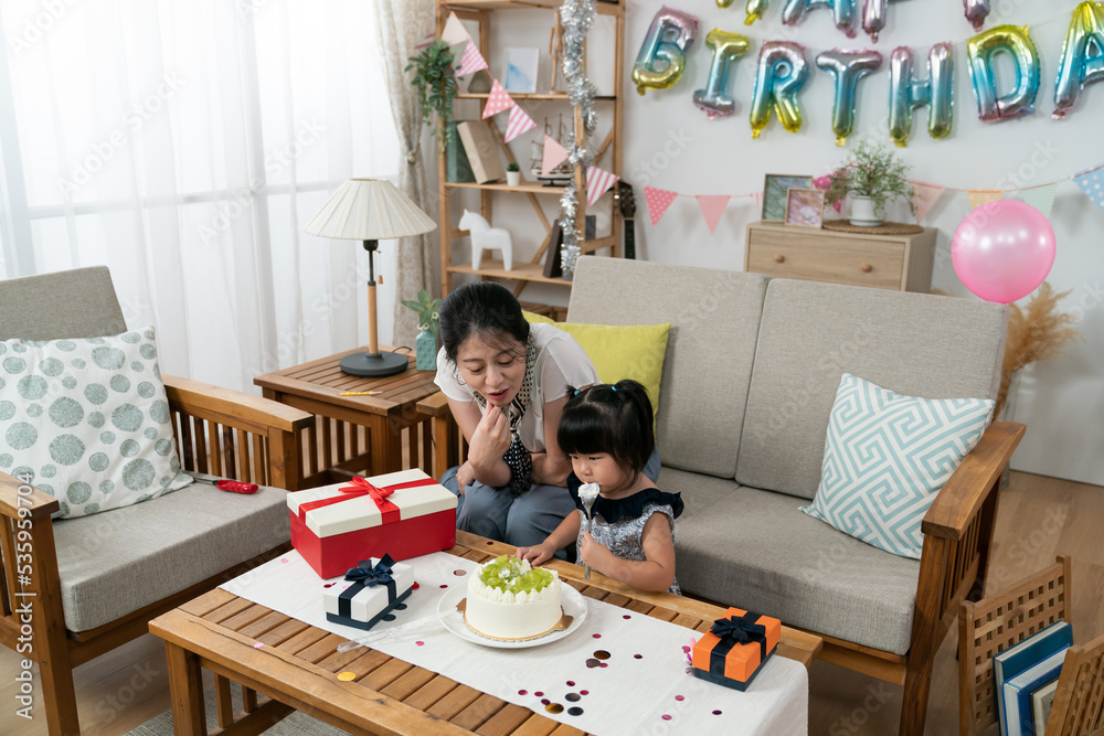 cute asian baby girl enjoying licking cream cake on the spoon in a modern home living room decorated for her second birthday celebration with her mother