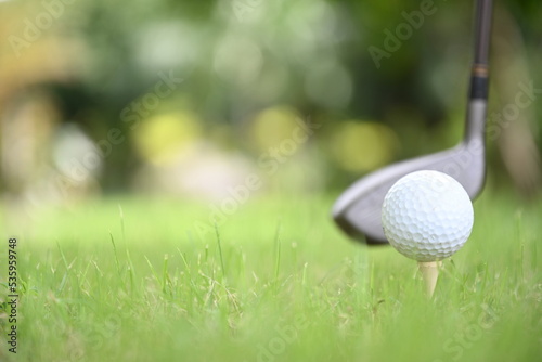 Blurred image of a white golf ball placed on a tee and golf club on a green lawn. People all over the world play golf during their health and leisure holidays. 